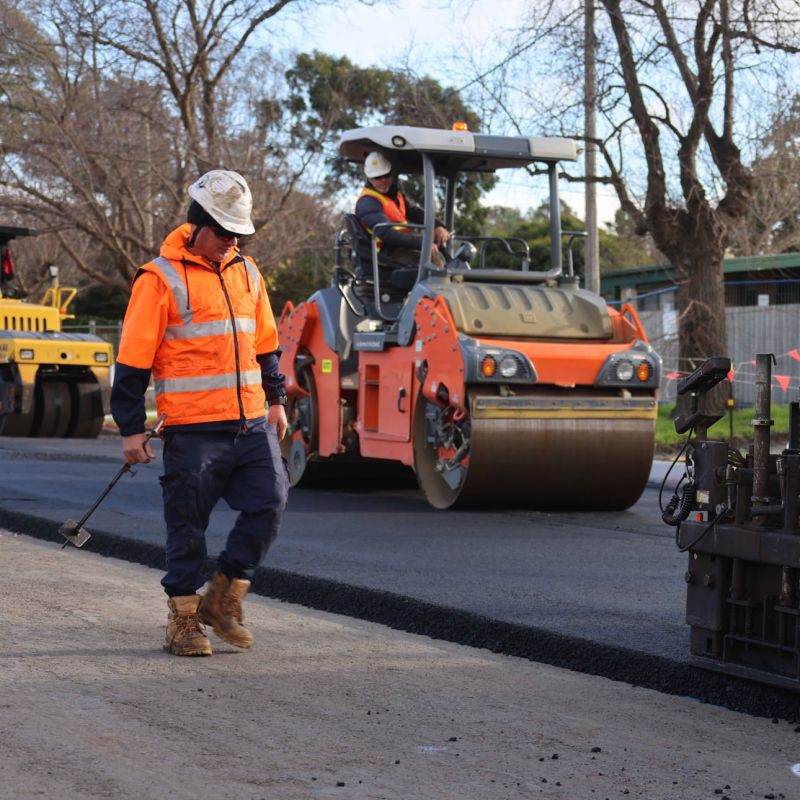 A Nexpave team member inspecting site quality while an asphalt roller operates in the background of a civil road project.